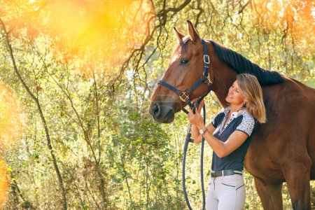 Foto de Contenido hembra ecuestre ajuste riendas en el caballo de castaño, mientras que de pie en el bosque en el día soleado en verano - Imagen libre de derechos
