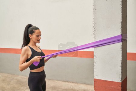 Téléchargez les photos : Vue latérale de l'athlète féminine déterminée dans les muscles de pompage de vêtements de sport à l'aide d'une bande de résistance violette enroulée autour de la colonne pendant l'entraînement dans un centre de fitness moderne - en image libre de droit