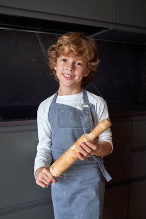 Téléchargez les photos : Joyeux garçon dans le tablier avec rouleau à pâtisserie en bois dans les mains regardant la caméra avec le sourire tout en se tenant dans la cuisine légère - en image libre de droit