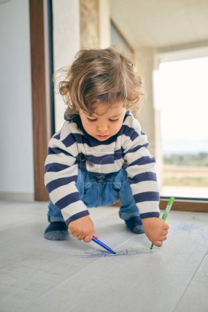 Photo for Adorable focused curly haired child in casual clothes sitting on haunches and drawing scribble on laminate floor with colorful markers near window in room - Royalty Free Image