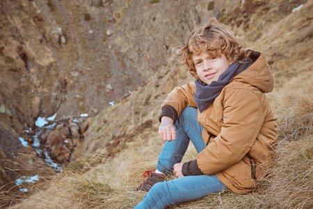 Photo for Side view of curly haired boy in warm outerwear sitting on dry grass on hillside and looking at camera - Royalty Free Image