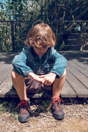 Téléchargez les photos : Corps complet de triste petit garçon en vêtements décontractés assis sur une jetée en bois avec la tête tombante dans la journée ensoleillée - en image libre de droit