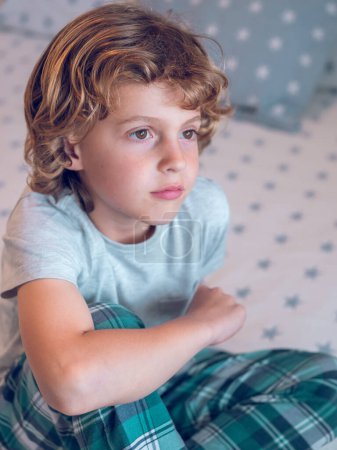 Photo for Cute preteen boy with curly blond hair in sleepwear sitting on bed and looking away and dreaming - Royalty Free Image