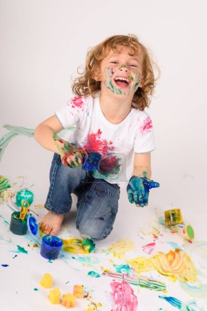 Foto de Cuerpo completo de niño alegre cubierto de pinturas de colores mirando a la cámara mientras está sentado en el suelo sucio sobre fondo blanco - Imagen libre de derechos