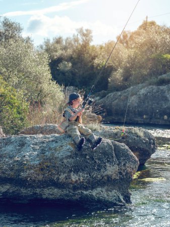 Photo for Boy in casual clothes looking at line of fishing rod while sitting on rough stone on sunny weekend day in countryside - Royalty Free Image