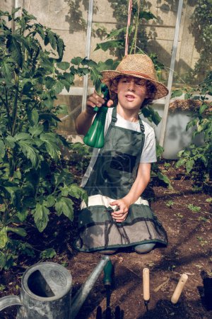 Photo for Kid in straw hat and apron sitting on ground with gardening tools and watering can and spraying growing plants - Royalty Free Image