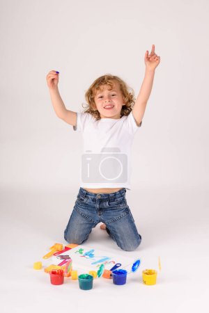 Photo for Full body of delighted boy in casual wear looking at camera with raised arms while sitting near set of paints on white background - Royalty Free Image