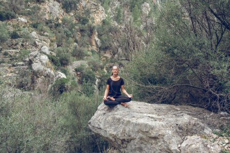 Téléchargez les photos : Pleine longueur de paisible jeune dame ethnique aux cheveux foncés en vêtements de sport méditant avec les yeux fermés et les mains mudra dans le yoga pose Lotus assis sur un rocher massif dans la vallée rocheuse - en image libre de droit