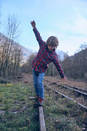 Téléchargez les photos : Corps complet de mignon garçon actif en chemise à carreaux équilibrage tout en se promenant sur le chemin de fer en métal dans la nature avec des arbres sans feuilles - en image libre de droit