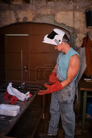 Foto de Vista lateral del soldador masculino con guantes protectores y casco mientras se prepara para el trabajo en taller gruñón - Imagen libre de derechos