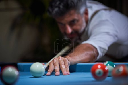 Foto de Hombre serio en camisa blanca de pie en la mesa de billar azul y preparándose para golpear la pelota con taco mientras juega al billar en la sala de estar oscura en casa - Imagen libre de derechos