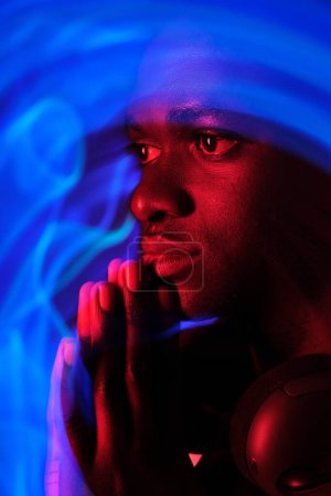 Téléchargez les photos : Vue latérale d'un homme afro-américain paisible avec des mains priantes sous une lumière fluorescente colorée - en image libre de droit