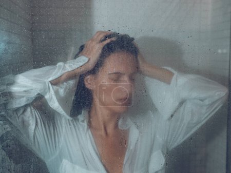 Foto de Triste dama empapada con los ojos cerrados en camisa blanca tocando el pelo detrás de cortina de ducha transparente - Imagen libre de derechos