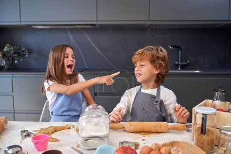 Téléchargez les photos : Fille riant au nez du frère sale avec de la pâte portant différents tabliers assis au comptoir de cuisine pâtisserie le week-end matin - en image libre de droit
