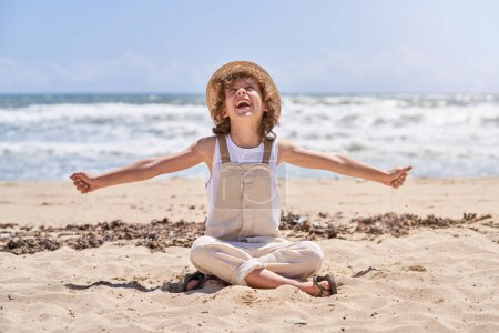 Foto de Nivel del suelo de cuerpo completo niño feliz en sombrero de paja y mono vertiendo arena mientras juega en la orilla del mar y mirando hacia arriba - Imagen libre de derechos