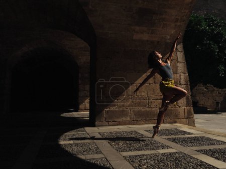 Photo for Full body side view of unrecognizable female performing ballet dance on paved street with shadow near shabby building in city - Royalty Free Image