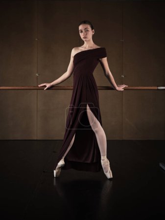 Foto de Cuerpo completo de elegante bailarina femenina en elegante vestido púrpura y zapatos de punta de pie en puntillas y apoyándose en la barra en el estudio de ballet y mirando a la cámara - Imagen libre de derechos
