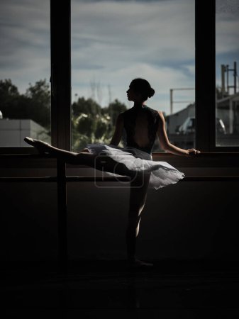 Téléchargez les photos : Vue arrière de la ballerine élancée debout à la barre de ballet et étirant la jambe levée pendant la répétition le soir - en image libre de droit