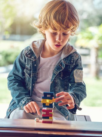 Téléchargez les photos : Concentré mignon garçon en jean veste jouer tour jeu seul à table dans la cour arrière sur ensoleillé journée d'été - en image libre de droit