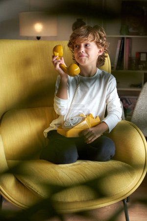 Téléchargez les photos : Corps complet d'enfant préadolescent mignon en tenue décontractée assis dans un fauteuil avec un téléphone filaire jaune vintage dans une pièce lumineuse avec une plante floue et détournant les yeux - en image libre de droit