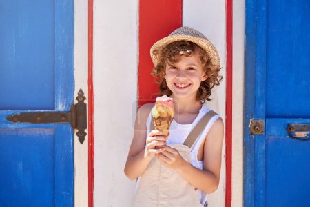 Foto de Feliz niño preadolescente rizado en ropa casual de pie cerca del edificio con puertas azules y comiendo delicioso cono de helado mientras mira a la cámara - Imagen libre de derechos