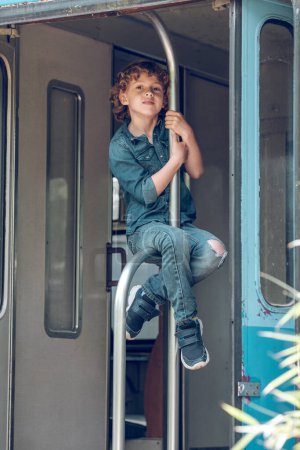 Photo for Full body of cheerful preteen boy climbing on metal handrail in doorway of shabby train and looking at camera - Royalty Free Image