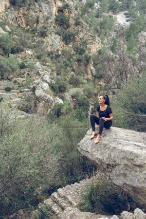 Téléchargez les photos : Voyageuse positive en vêtements décontractés assise sur le rocher et admirant les montagnes tout en voyageant dans la campagne de Majorque, Espagne - en image libre de droit