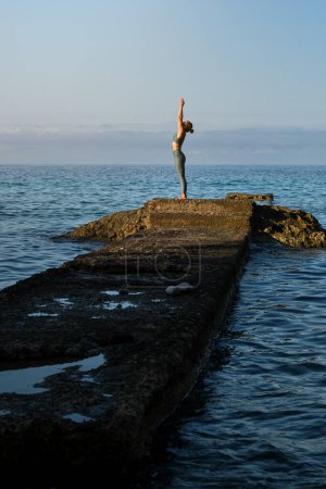 Téléchargez les photos : Vue latérale d'une femme en forme en tenue de sport debout sur une côte rocheuse pratiquant le yoga près d'agiter la mer contre le ciel - en image libre de droit
