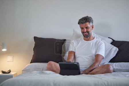 Photo for Mature male entrepreneur in sleepwear smiling and looking at screen of tablet while working on project remotely in bedroom at home - Royalty Free Image