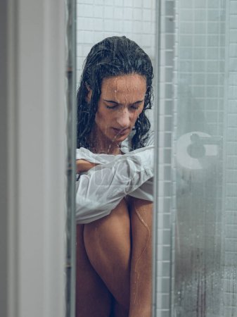 Foto de Mujer hispana agotada en camisa blanca sentada en el suelo de la cabina de ducha bajo el agua salpicada con los ojos cerrados - Imagen libre de derechos