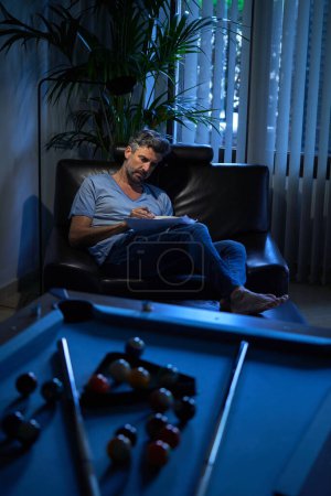 Photo for Full body of serious businessman in casual clothes writing notes while sitting on chair working late in recreation room at office - Royalty Free Image
