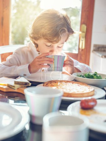 Foto de Adorable chico con el pelo rizado desayunando mientras está sentado en la mesa con deliciosa comida cerca de la puerta en el comedor - Imagen libre de derechos