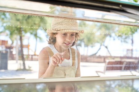 Photo for Through glass of cheerful boy in straw hat standing near showcase and choosing ice cream in park - Royalty Free Image