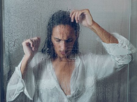 Téléchargez les photos : À travers le verre de femme bouleversée avec les cheveux mouillés debout près de la porte brumeuse de la cabine de douche avec des gouttes d'eau dans la salle de bain - en image libre de droit