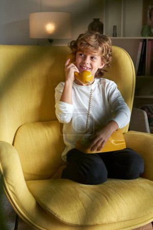 Téléchargez les photos : Corps complet de garçon positif avec des cheveux bouclés parlant sur le téléphone fixe rétro dans le fauteuil dans la pièce lumineuse avec plante floue - en image libre de droit