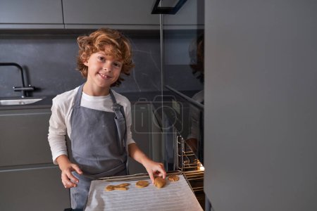 Foto de Niño sonriente en delantal gris poniendo galletas de diferentes formas en la cocina estufa y mirando a la cámara - Imagen libre de derechos