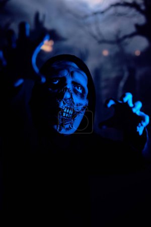 Photo for Awful undead monster stretching out arms and limping towards camera under blue neon light at night on dark cemetery - Royalty Free Image