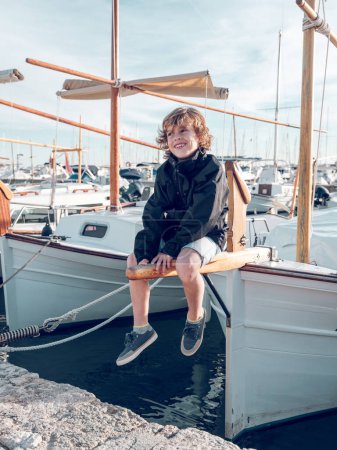 Téléchargez les photos : Joyeux garçon en vêtements décontractés souriant et assis sur un voilier lors d'une journée d'été ensoleillée dans le port de Pollensa à Majorque, Espagne - en image libre de droit