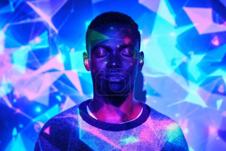 Téléchargez les photos : Homme afro-américain tranquille avec les yeux fermés debout dans l'éclairage au néon dans un studio lumineux - en image libre de droit