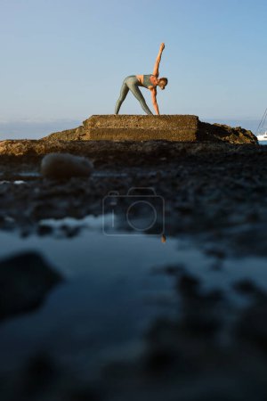 Photo for Distant anonymous female in sportswear performing yoga in Triangle pose on rough rocky cliff against cloudless blue sky - Royalty Free Image