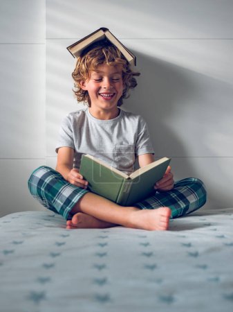 Foto de Cuerpo completo de niño sonriente en ropa de dormir sentado en la cama con las piernas cruzadas y leyendo una historia interesante con la cabeza cubierta de libro - Imagen libre de derechos
