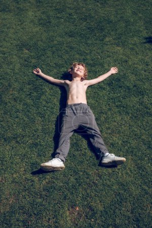 Téléchargez les photos : D'en haut corps plein de petit enfant satisfait avec torse nu couché sur l'herbe verte et étirant les bras et les jambes - en image libre de droit