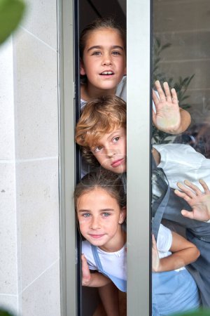 Téléchargez les photos : Positif mignon curieux enfants en vêtements décontractés debout près du mur blanc et appuyé sur la porte en verre tout en regardant à l'extérieur - en image libre de droit