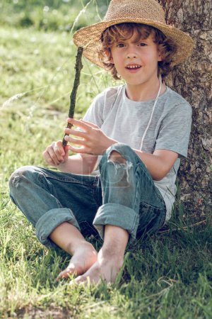 Photo for Full body of preteen boy in wicker hat and dirty clothes sitting on grass under tree with branch and looking at camera - Royalty Free Image
