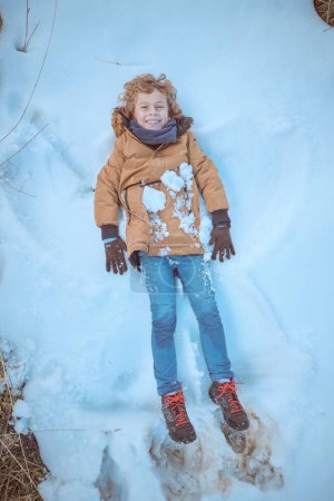 Téléchargez les photos : Vue du dessus corps complet de garçon ludique en vêtements de dessus sans chapeau couché sur la neige et faire ange tout en s'amusant dans la nature hivernale - en image libre de droit