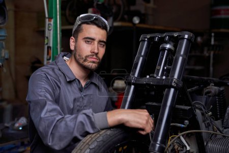 Photo for Serious bearded male mechanic in uniform sitting near motorbike with hand on wheel and looking at camera while working in professional workshop - Royalty Free Image
