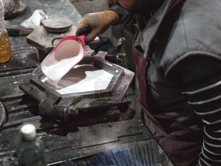 Téléchargez les photos : Angle élevé de culture artisan masculin anonyme dans des gants de protection en utilisant une cuillère en plastique tout en versant de la céramique blanche dans un moule en métal pour produire des carreaux de céramique à l'usine - en image libre de droit