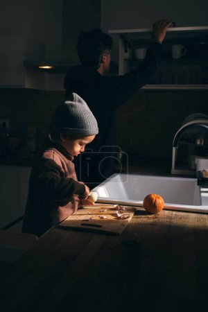 Foto de Vista lateral de lindo niño atento en ropa elegante y sombrero pelando mandarinas frescas mientras está de pie en la mesa cerca de padre irreconocible en busca de ingredientes en la cocina - Imagen libre de derechos