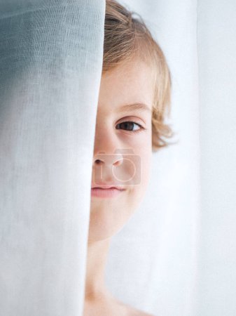 Foto de Crop boy con el pelo rubio asomándose por detrás de la cortina y mirando a la cámara - Imagen libre de derechos