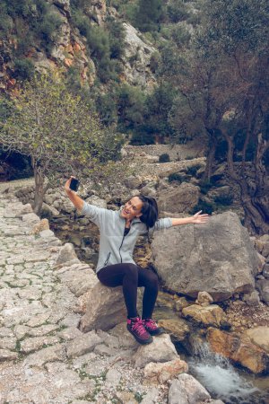 Téléchargez les photos : Corps entier de jeune voyageuse ethnique excitée avec de longs cheveux foncés en vêtements décontractés criant joyeusement tout en prenant selfie sur téléphone portable assis sur le rocher au bord de la rivière pendant la randonnée - en image libre de droit
